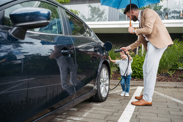 Mature man and his little son charging their electric car on a rainy day, standing under umbrella.