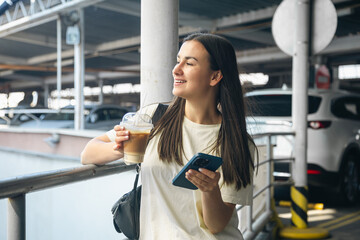 A young woman with coffee and smartphone in the parking lot.