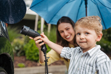 Close up of smiling mother and her little son charging their electric car on a rainy day, standing...