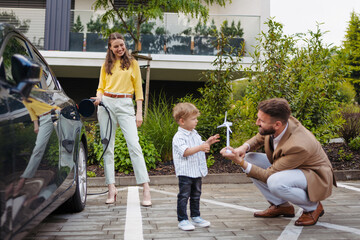 Happy family standing beside their car and charging electric car on the street.