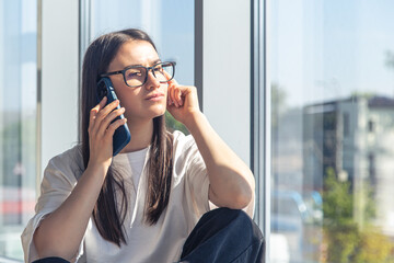 A young woman in glasses is talking on a smartphone near the window.
