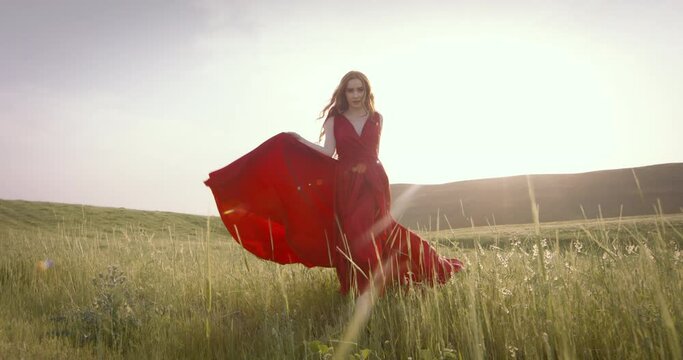 Caucasian woman model in fashionable red dress confidently walks across field at sunset. Style, fashion, latest fashion trends, fashion image. Confident gait of brave and daring woman in red dress