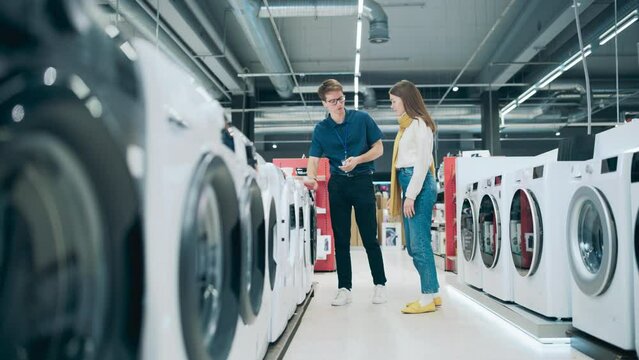 Young Female Customer Engages with Home Electronics Store Associate while Selecting a Washing Machine. Stylish Woman Seeks High-Quality Appliance. Shopper Evaluating Modern Laundry Solutions