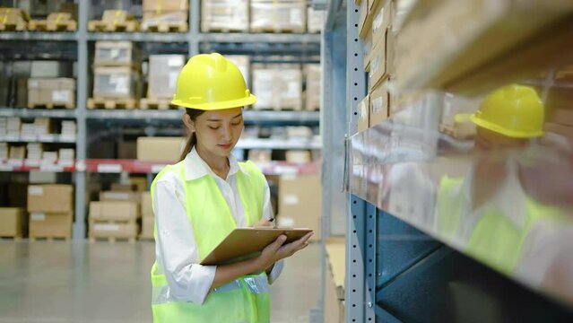 Asian woman warehouse worker in uniform doing stocktaking of products management in cardboard box on shelves in warehouse using clipboard file and pen and looking camera, logistic and business export