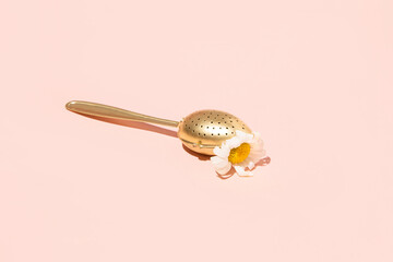 Golden tea infuser spoon, daisy flower trapped in it, creative aesthetic layout, pastel pink background.