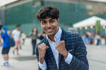 A stylish guy in a blue checkered suit, smiling with arms up in a fighting position leaning forward while standing outside, in the city. Building, people and tents are shown in the background.