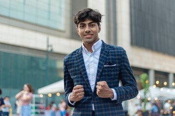A stylish guy in a blue checkered suit, smiling at the camera, showing two boxing fist hand gesture while standing outside, in the city. Building, people and tents are shown in the background.