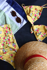 Yellow floral bikini, straw hat, striped towel and sunglasses on dark background. Flat lay.
