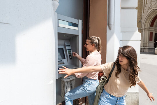 Two young female tourists, world travelers, one withdraws money with a credit card from an ATM, the other guards her from thieves and fraudsters