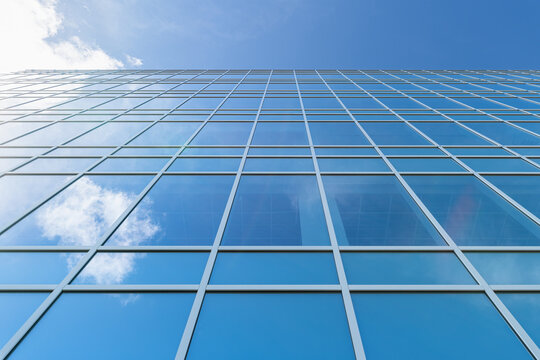 A low angle photograph of the side of a tall building made from glass as it reflects the blue cloudy sky