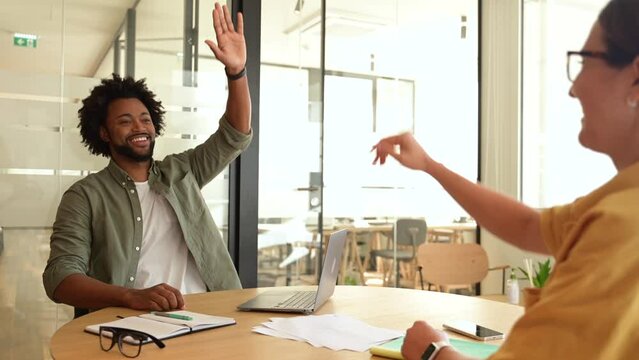 Two young happy diverse colleague sitting at desk and taking high five while working together in coworking space, man and woman celebrating successful project completion or good deal