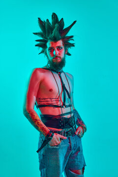 Shirtless young man, rock musician punk with crazy hairdo and makeup posing against blue studio background in neon light. Concept of music, lifestyle, subculture, art, youth, human emotions