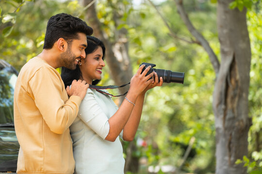 Happy young Indian woman with husband taking photos in front of car while traveling - concept of hobbies,togetherness and holidays.
