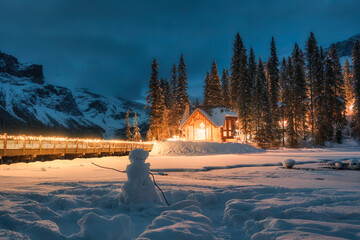 Snowy Emerald lake with wooden lodge glowing in pine forest and snowman in the night at Canada