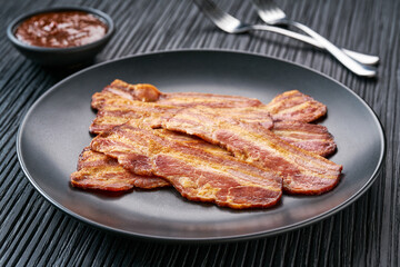 fried bacon with bbq sauce in a dish on a black table. Fried crunchy streaky bacon pieces in a black plate.