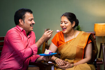 Happy husband feeding cake to wife while sitting on sofa at home - concept of wedding anniversary...