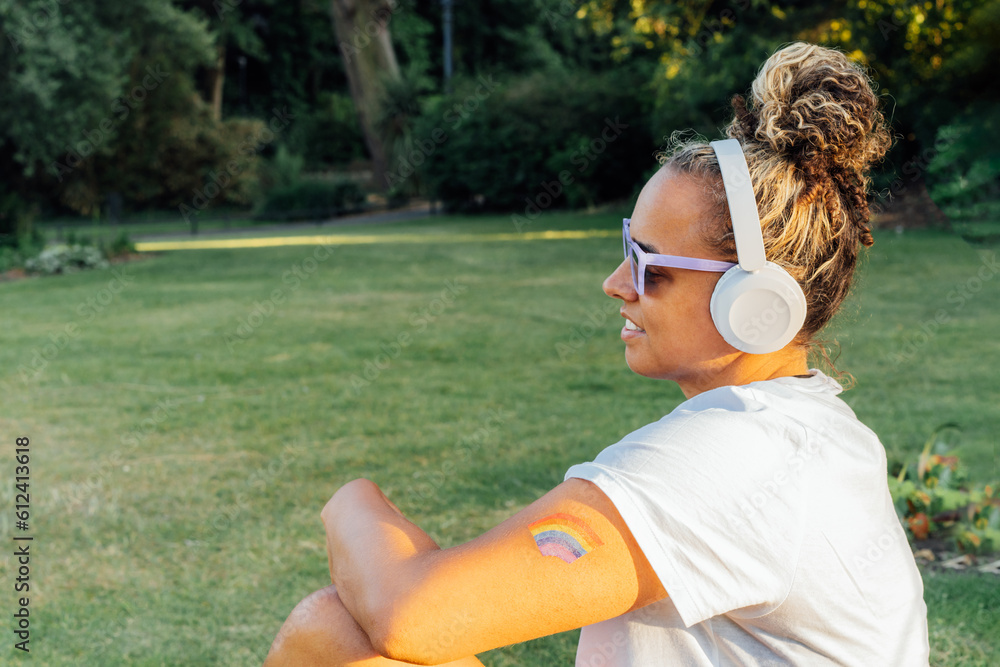 Wall mural Side view young woman with rainbow tattoo, symbol of LGBTQ social community listening music with wireless headphones while relax in park. Personal Coming out, free lifestyle. Celebrating Pride month.