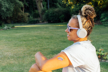 Side view young woman with rainbow tattoo, symbol of LGBTQ social community listening music with wireless headphones while relax in park. Personal Coming out, free lifestyle. Celebrating Pride month. - Powered by Adobe