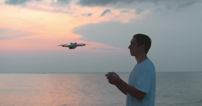 Silhouette of man traveler control drone quadcopter to take video or photo on seascape background in the evening while traveling. Man drone pilot with controller launch copter aircraft in sky at sea