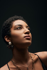 Fototapeta premium Portrait of elegant and short haired african american woman with evening makeup and golden accessories looking away and standing isolated on black, high fashion and evening look