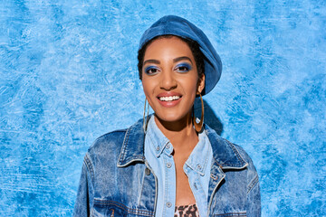 Portrait of positive young african american woman with bold makeup in beret and denim jacket looking at camera on blue textured background, stylish denim attire