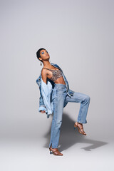 Full length of fashionable african american woman with bold makeup posing in top with animal print, denim jacket and jeans while standing on grey background, denim fashion concept