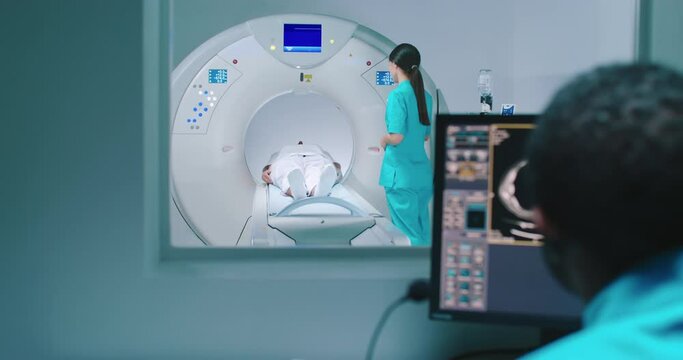 African American doctor observes process of MRI examination. Nurse send patient to MRI capsule on moving table. Magnetic resonance imaging of brain. Room of examinaning with medical equipment.