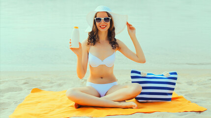 Summer vacation, happy smiling young woman sunbathing with suntan cream wearing bikini and straw hat lying on sand on the beach