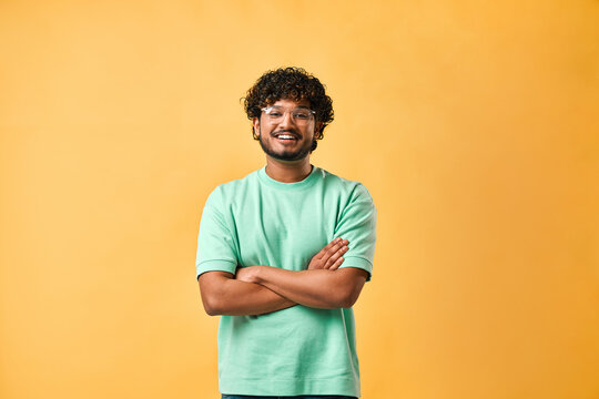 Image of a handsome young man in glasses and a turquoise t-shirt standing on a yellow background with crossed arms and smiling.