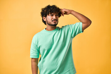 Portrait of a handsome young man in a turquoise t-shirt on a yellow background, scratching his head and looking very pensively to the side. The emotion of doubt, reflection and confusion. - obrazy, fototapety, plakaty