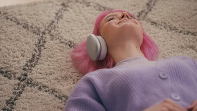 Relaxed woman with pink hair lying down on floor, listening to music in headset