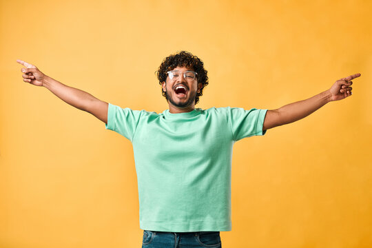 Victory celebration. An emotional handsome young man with curly hair in a turquoise T-shirt on a yellow background points his hands to the side and shouts. The emotion of happiness, joy, laughter.