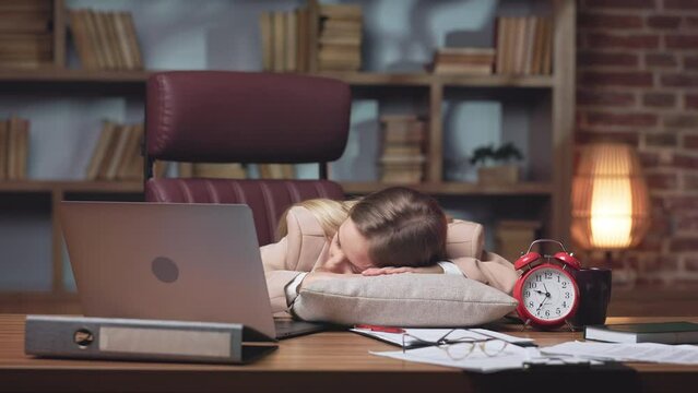 Tired female office employee fluffing up pillow and falling asleep. Young caucasian woman getting exhausted after intense working day with computer and taking short nap in elegant office place.