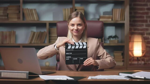 Portrait of long-haired blonde actress sitting at table with laptop and holding black clapperboard in hands. Beautiful model playing at casting audition. Concept of cinema and film industry.
