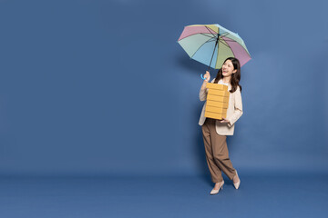 Happy Young Asian woman standing and holding package parcel box and rainbow umbrella isolated on blue background, Delivery courier and shipping service in rainy season concept