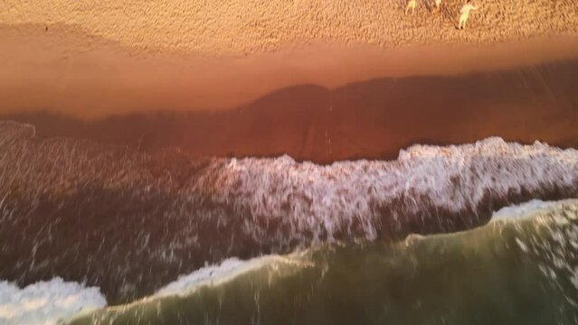 Drone shot over sea waves moving to people on the beach rasing on green rocky mountains at sunset