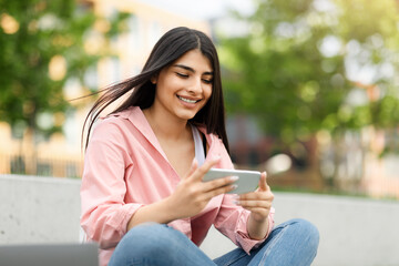 Obraz premium Smiling teen hispanic lady messaging or playing on smartphone while relaxing in park outdoors, free space