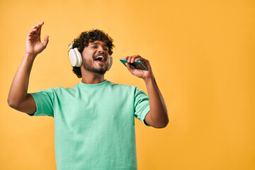 Portrait of handsome curly Indian man in turquoise t-shirt and singing with phone in wireless...