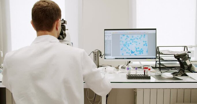 Male doctor using microscope in laboratory. Man in lab coat and latex gloves using microscope to examine sample during work in clinic in daytime. Microscope for blood examination at the clinic.