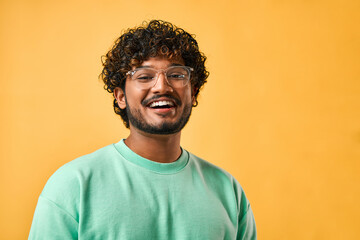 Close-up portrait of a handsome curly-haired Indian man in a turquoise t-shirt and glasses laughing and looking at the camera. - obrazy, fototapety, plakaty