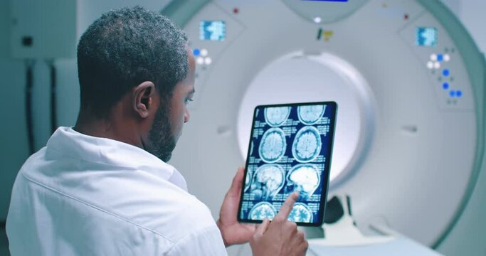 African American doctor examine scan attentively. Medical worker shooted from back. Doctor looks at MRI scan. Multicultural doctor is scrolling screen with magnetic resonance imaging scans of brain.