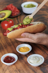 Fototapeta na wymiar Completo italiano: vertical. Adding palta, with wooden spoon, on tomato and vianesa on hot dog bun, one hand, tomato, avocado, mayonnaise and mustard on a wooden table. Typical Chilean food concept