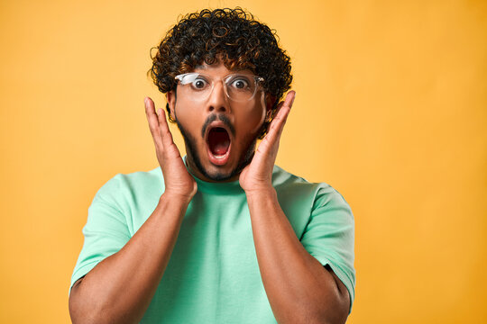Close-up portrait of a handsome Indian man with curly hair in a turquoise t-shirt and glasses with big eyes and open mouth standing on a yellow background and expressing the emotion of shock, surprise