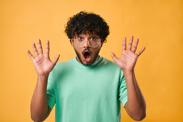 Close-up portrait of handsome Indian man in turquoise t-shirt and glasses with big eyes and open mouth standing on yellow background and expressing emotion of shock, surprise and showing hands.
