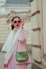 Fashionable elegant confident woman wearing trendy pink sunglasses, suit blazer, white silk scarf, trousers, with green faux leather shoulder bag, posing in street. Outdoor fashion portrait - 612457656