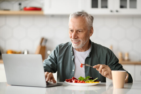 Smiling Senior Man Using Laptop And Having Breakfast In Kitchen