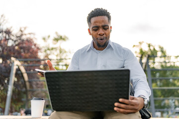 A well-dressed young Black man sits on a riverside promenade, engaged in a lively discussion with colleagues through his laptop. 