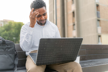 A tired young Black man, dressed in a business attire, sits on a bench, checking his personal...