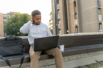 A tired young Black man, dressed in a business attire, sits on a bench, checking his personal...