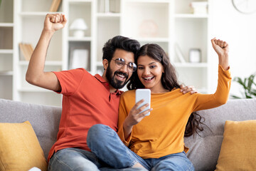 Joyful Indian Couple Celebrating Success While Looking At Smartphone Screen At Home